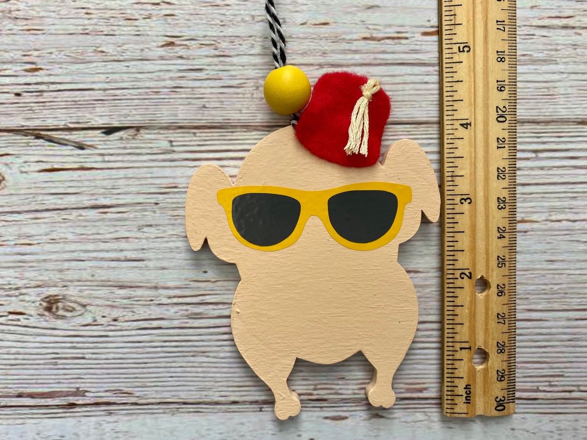 A wooden turkey-shaped ornament or plaque with a red felt hat, yellow sunglasses, and a black-and-white hanging cord, shown on a whitewashed wood surface next to a wooden ruler for scale.