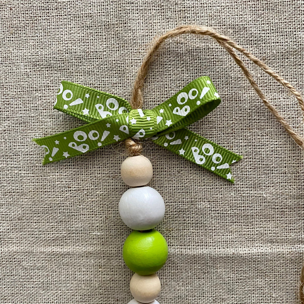 A decorative hanging ornament with a green ribbon bow printed with white 'boo' text, tied to natural jute string. Below the bow are four wooden beads in light beige, white, bright green, and light beige, arranged on a textured beige fabric background.