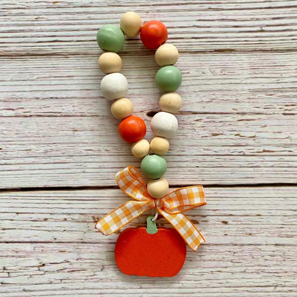 A fall-themed wooden bead loop with orange, green, beige, and white beads, finished with an orange pumpkin charm and an orange and white gingham ribbon bow, displayed on a rustic white-washed wooden background.