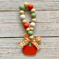 A fall-themed wooden bead loop with orange, green, beige, and white beads, finished with an orange pumpkin charm and an orange and white gingham ribbon bow, displayed on a rustic white-washed wooden background.