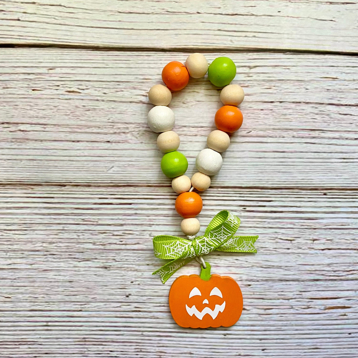 A beaded loop garland with orange, green, and natural wood beads, decorated with a green spiderweb ribbon and a carved orange pumpkin charm, displayed on a rustic white-washed wooden background.