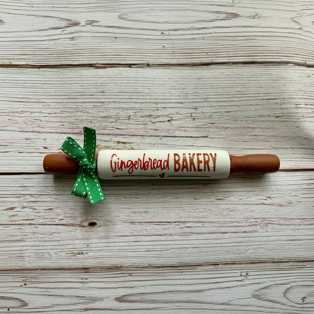 A gingerbread-themed rolling pin with 'Gingerbread BAKERY' written on it, featuring brown wooden ends, a white center, and a green ribbon bow, set against a rustic white wood background.