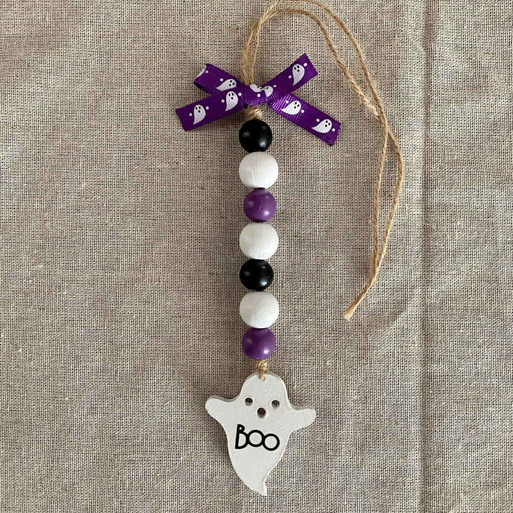 A Halloween-themed beaded ornament with purple, white, and black beads, a purple ribbon bow, and a white ghost-shaped charm with 'boo' written on it, displayed on a beige linen background.
