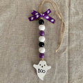A Halloween-themed beaded ornament with purple, white, and black beads, a purple ribbon bow, and a white ghost-shaped charm with 'boo' written on it, displayed on a beige linen background.