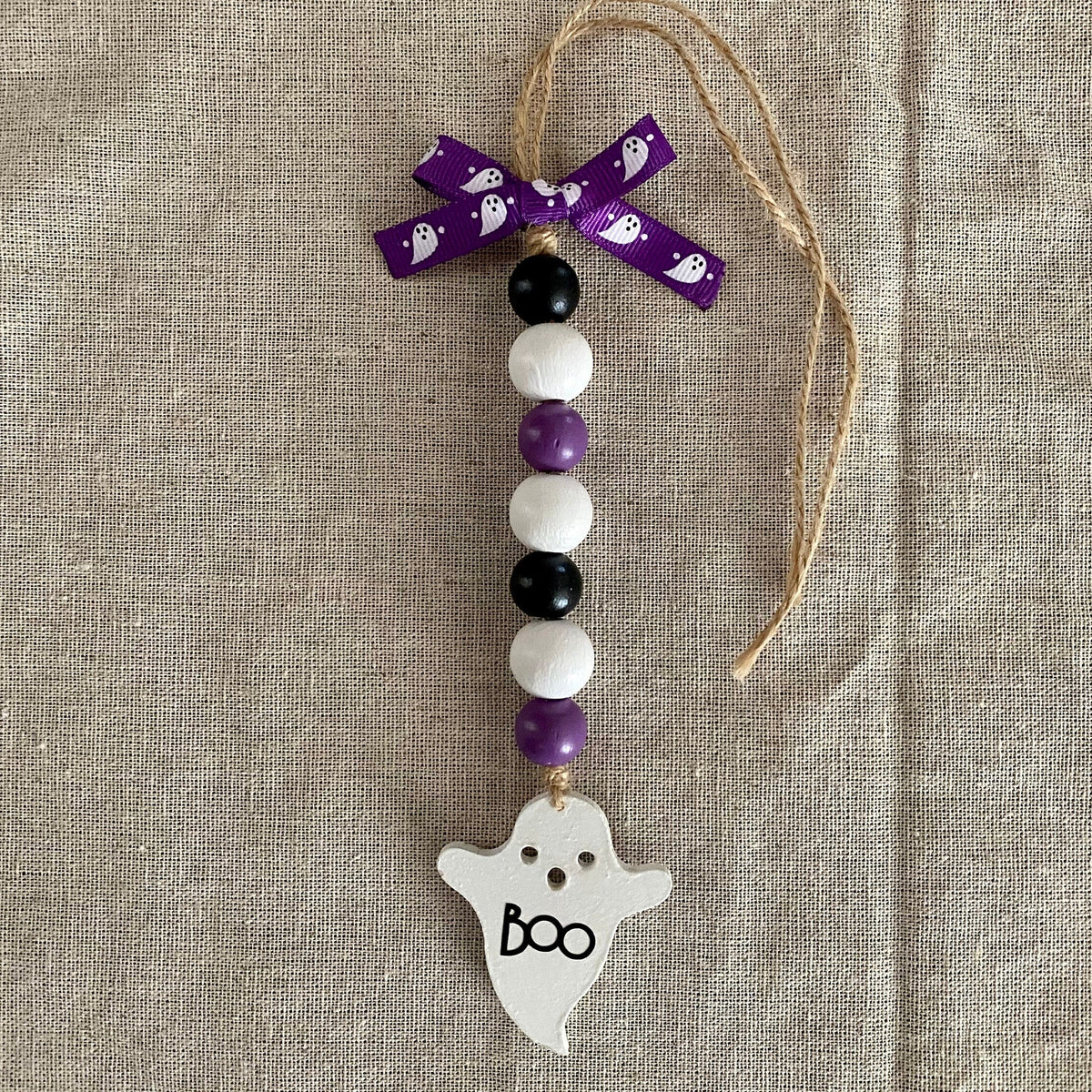 A Halloween-themed beaded ornament with purple, white, and black beads, a purple ribbon bow, and a white ghost-shaped charm with 'boo' written on it, displayed on a beige linen background.
