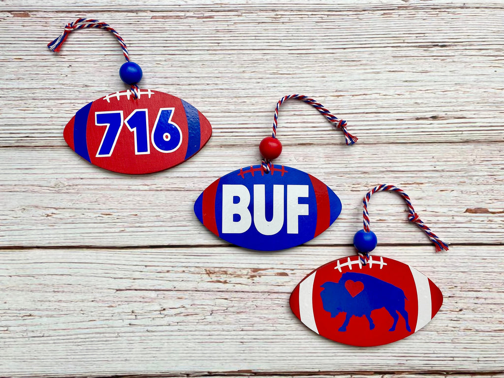 Three wooden football-shaped ornaments in red, white, and blue are arranged on a whitewashed wood background. One ornament shows the number 716, another says BUF, and the third features a buffalo silhouette.