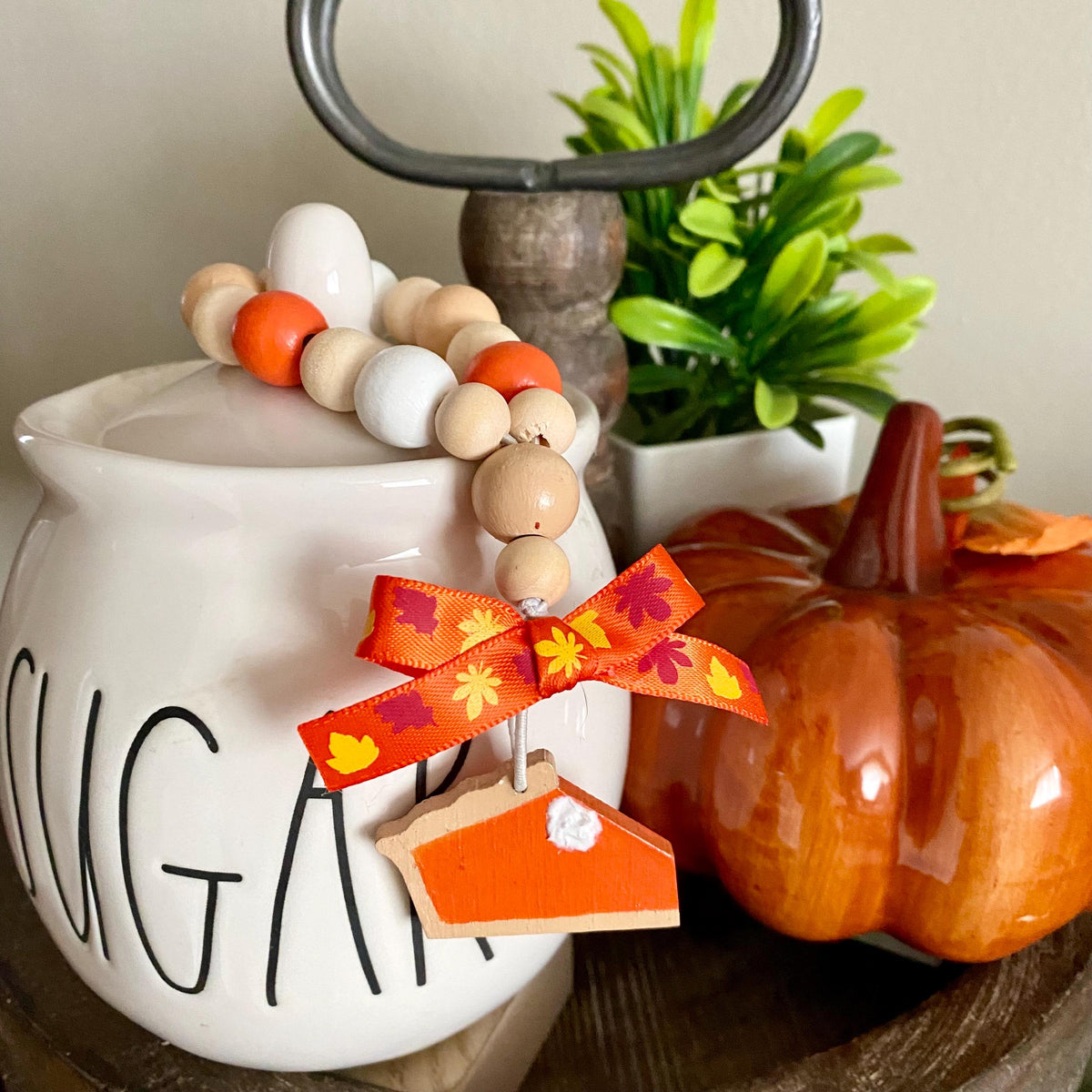A decorative beaded garland with orange, white, and natural wood beads, an orange and yellow autumn leaf ribbon, and a small house-shaped pie cutout charm, draped over a white ceramic sugar jar in a styled fall scene.