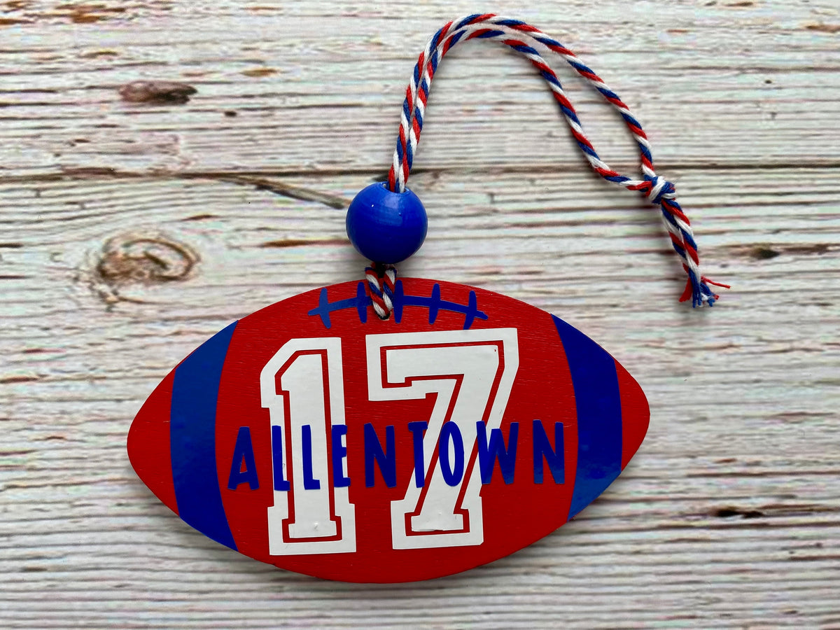 A football-shaped wooden ornament with the name 'ALLENTOWN' and number '17' in blue and white, hanging from a red, white, and blue rope on a rustic wood background.
