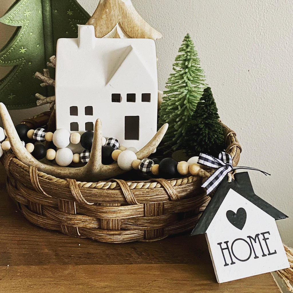 A rustic home decor arrangement in a woven basket, featuring a white ceramic house ornament, a small green tree, black-and-white beads, antler garland, and a hanging white house tag with the word HOME and a heart.
