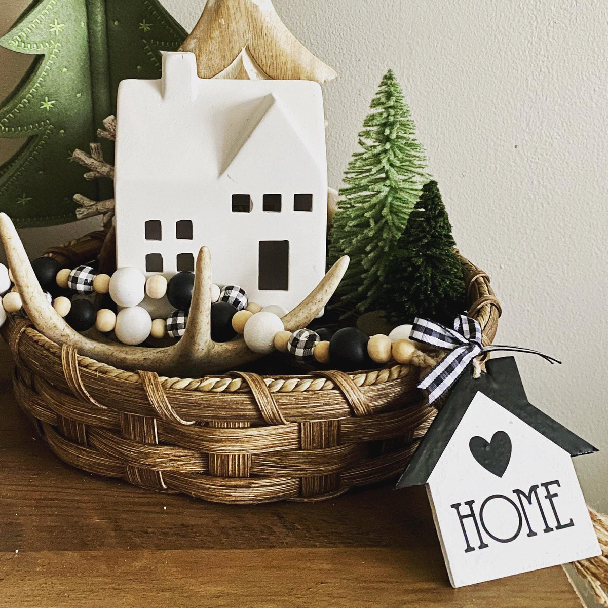 A rustic home decor arrangement in a woven basket, featuring a white ceramic house ornament, a small green tree, black-and-white beads, antler garland, and a hanging white house tag with the word HOME and a heart.