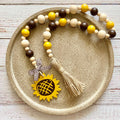 A wooden bead garland with yellow, cream, and dark brown beads, a beige jute tassel, and a yellow sunflower charm, displayed on a round textured gold tray over a white wood background.