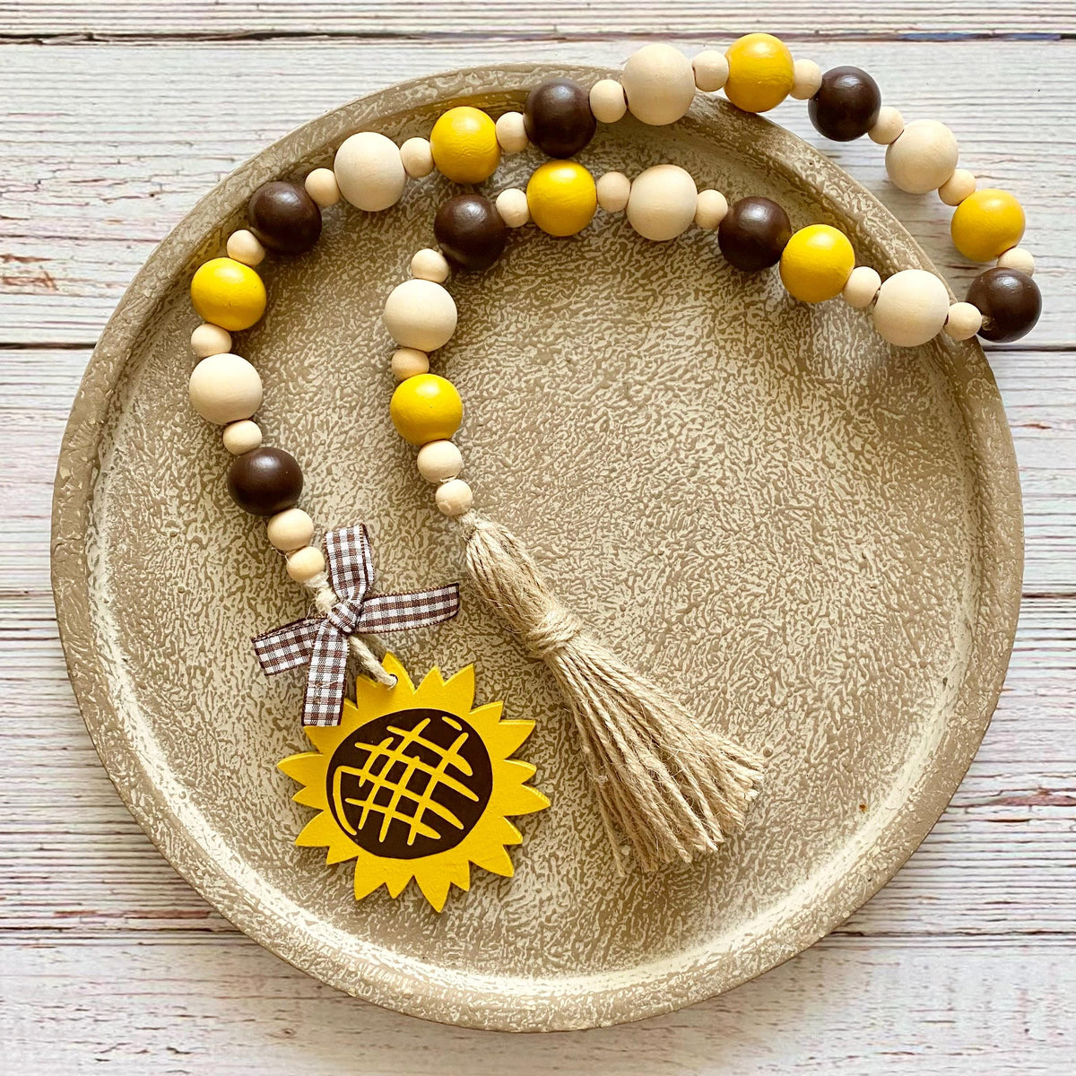 A wooden bead garland with yellow, cream, and dark brown beads, a beige jute tassel, and a yellow sunflower charm, displayed on a round textured gold tray over a white wood background.