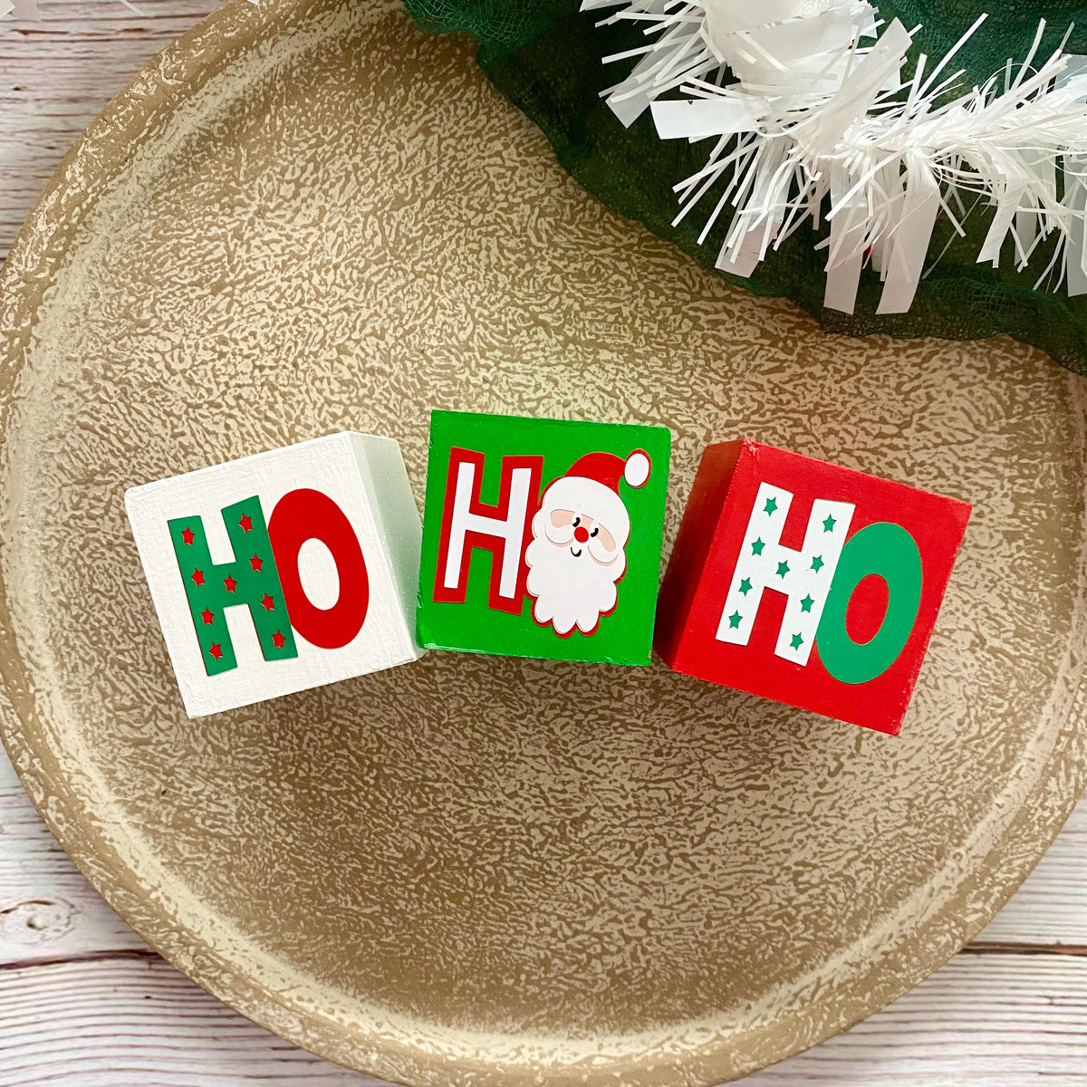 Three wooden Christmas letter blocks spelling 'HO HO HO' with festive holiday designs, arranged on a textured gold plate.
