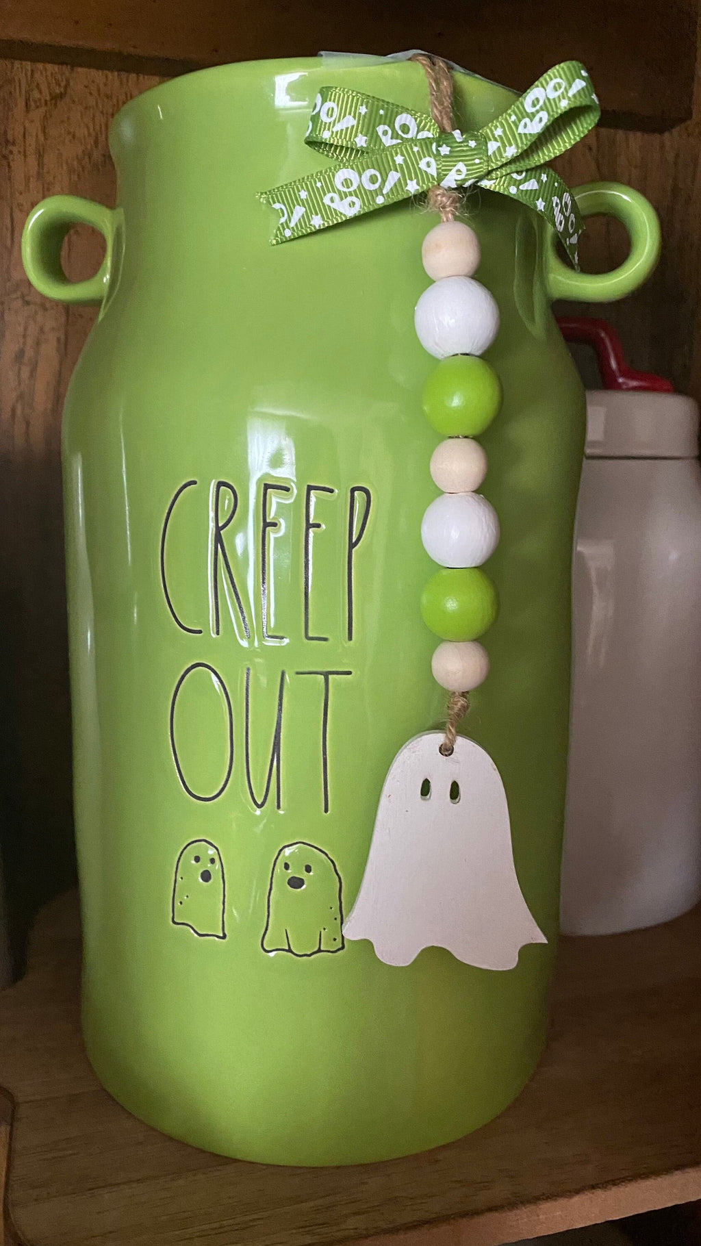 A bright green ceramic milk canister with 'CREEP OUT' and ghost illustrations, decorated with a green patterned ribbon bow and a beaded hanging ornament with a white ghost.