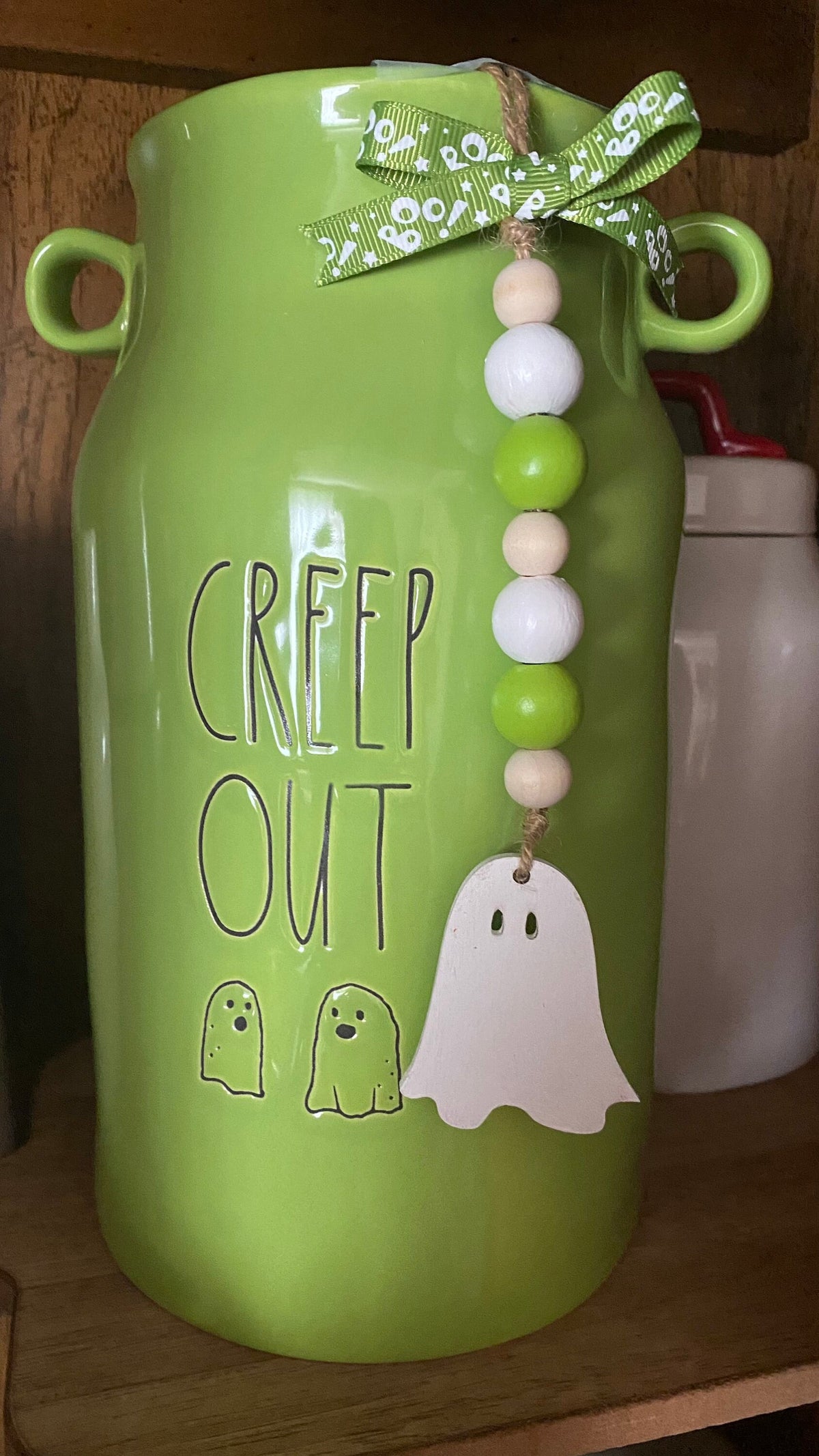 A bright green ceramic milk canister with 'CREEP OUT' and ghost illustrations, decorated with a green patterned ribbon bow and a beaded hanging ornament with a white ghost.