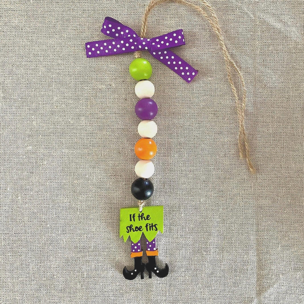 A Halloween-themed beaded ornament featuring purple, green, white, purple, orange, black, and white beads, with a purple polka-dot ribbon bow at the top and a dangling green sign that reads 'If the shoe fits' above black witch legs. The ornament is displayed on a textured beige linen background.