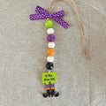 A Halloween-themed beaded ornament featuring purple, green, white, purple, orange, black, and white beads, with a purple polka-dot ribbon bow at the top and a dangling green sign that reads 'If the shoe fits' above black witch legs. The ornament is displayed on a textured beige linen background.