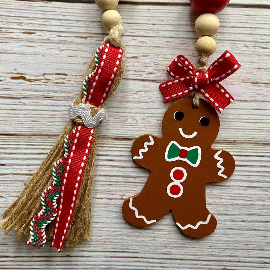 A close-up of a Christmas-themed beaded garland on a whitewashed wood background. The garland features light wood beads, a red and white striped ribbon tassel with a silver bow accent, and a brown wooden gingerbread man ornament with white icing details, a green bow tie, and red buttons.