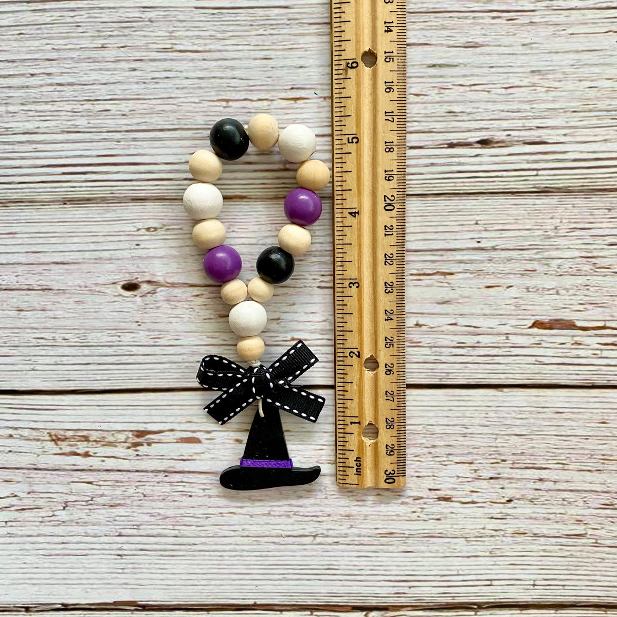 A beaded garland with purple, black, and natural wood beads, finished with a black witch hat charm and black-and-white polka dot bow, shown next to a wooden ruler on a whitewashed wood plank background.