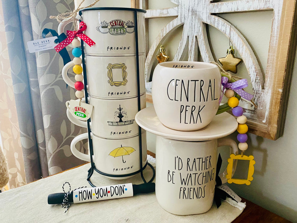 A set of ceramic mugs and matching canisters styled with 'Central Perk' themed graphics, including the phrases 'Central Perk', 'I'D Rather BE WATCHING FRIENDS', and 'HOW -YOU -DO -IN'. The set includes a black metal tiered canister, a stack of two mugs on a saucer, and colorful bead garlands in a rustic home decor setting.