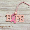 A pink and blush wooden letter ornament spelling 'HOPE' with a pink awareness ribbon through the center, hanging from a pink and white string on a whitewashed wood background.