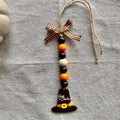 A handmade Thanksgiving ornament featuring a wooden pilgrim hat charm with 'Just Thanks' written on it, hanging from a brown and white gingham ribbon bow with orange, brown, and cream wooden beads, displayed on a textured gray linen fabric background.