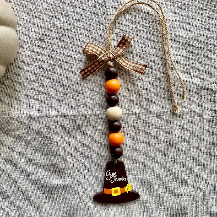A handmade Thanksgiving ornament featuring a wooden pilgrim hat charm with 'Just Thanks' written on it, hanging from a brown and white gingham ribbon bow with orange, brown, and cream wooden beads, displayed on a textured gray linen fabric background.