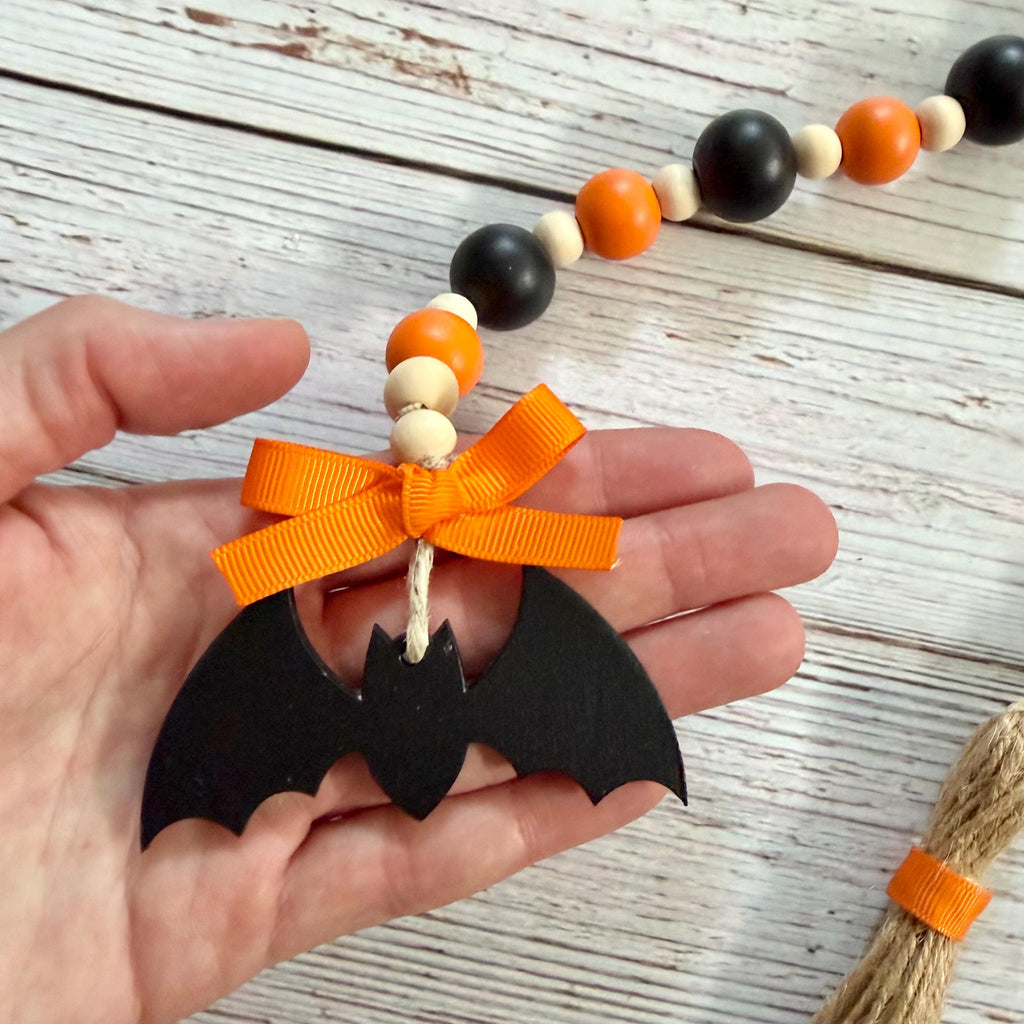 A hand holds a black bat-shaped ornament with an orange ribbon bow, attached to a beaded garland with orange, black, cream, and natural wood beads, on a whitewashed wood background.