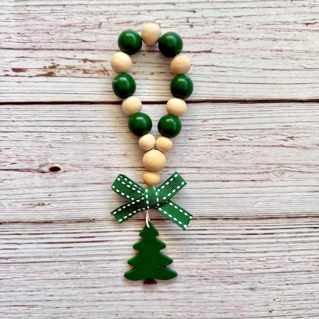 A wooden bead garland ornament with alternating dark green and natural beige wooden beads, a green patterned ribbon bow, and a green Christmas tree charm, displayed on a rustic whitewashed wood background.