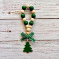 A wooden bead garland ornament with alternating dark green and natural beige wooden beads, a green patterned ribbon bow, and a green Christmas tree charm, displayed on a rustic whitewashed wood background.