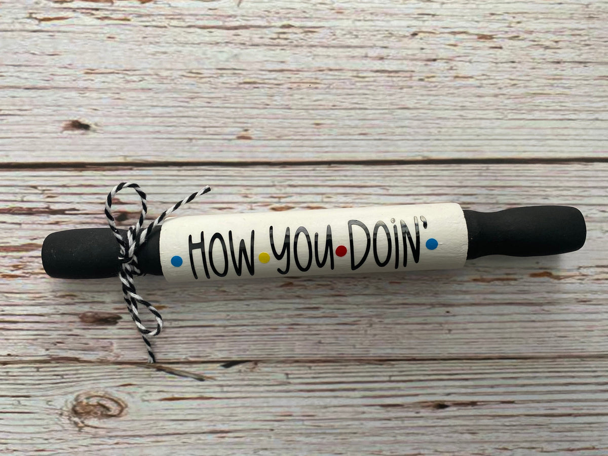A novelty rolling pin with a white body, black handles, and the phrase 'HOW YOU DOIN'' printed on it in black with small colored dots, tied with a black-and-white string, on a rustic white wood background.