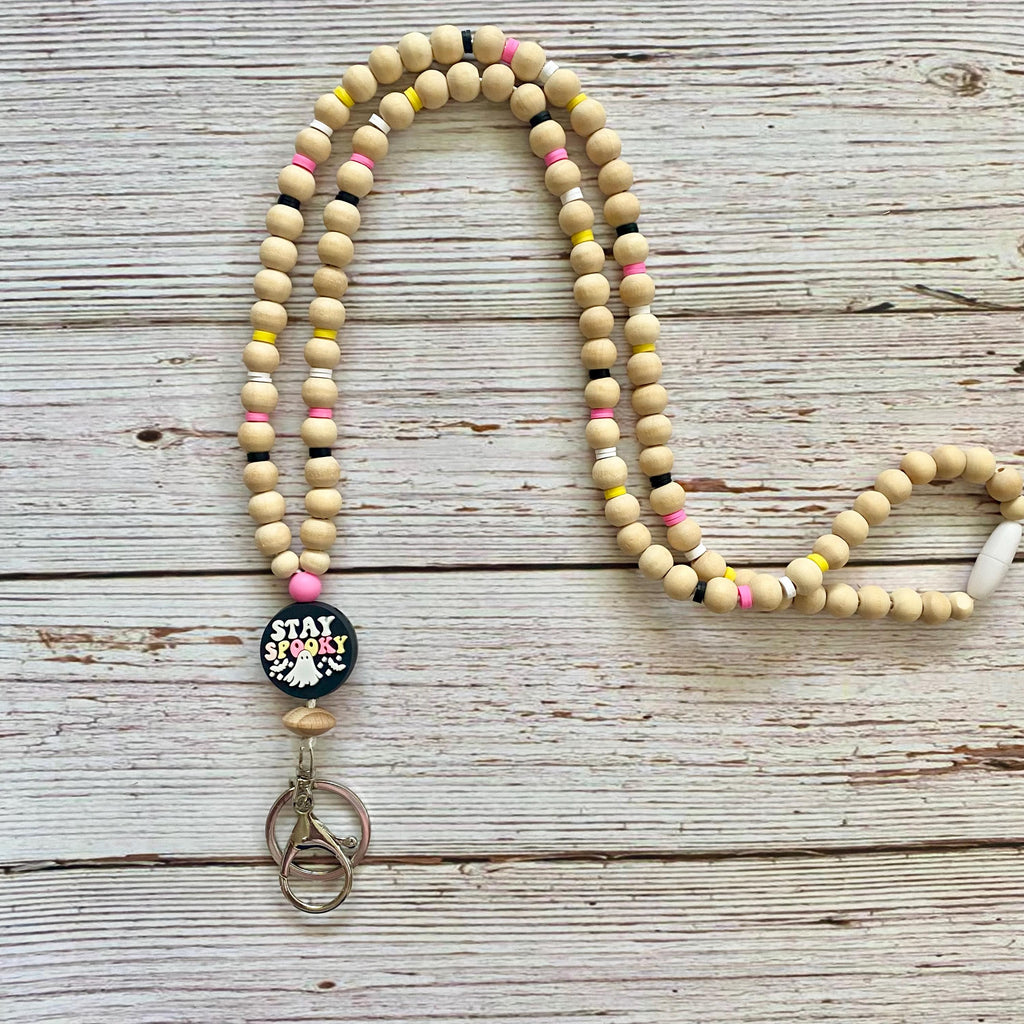 A beaded necklace with natural wood beads, small pink, yellow, and black accent beads, and a round black button charm that reads 'STAY spooky!' with a metal keychain attached, displayed on a rustic white-washed wooden background.