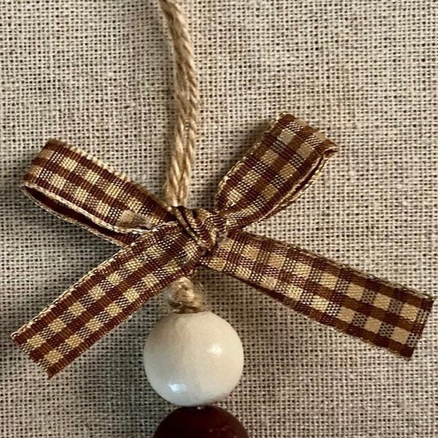 A rustic Christmas ornament featuring a brown and beige gingham ribbon bow, a white wooden bead, and a dark brown wooden bead, hanging against a textured beige burlap background.