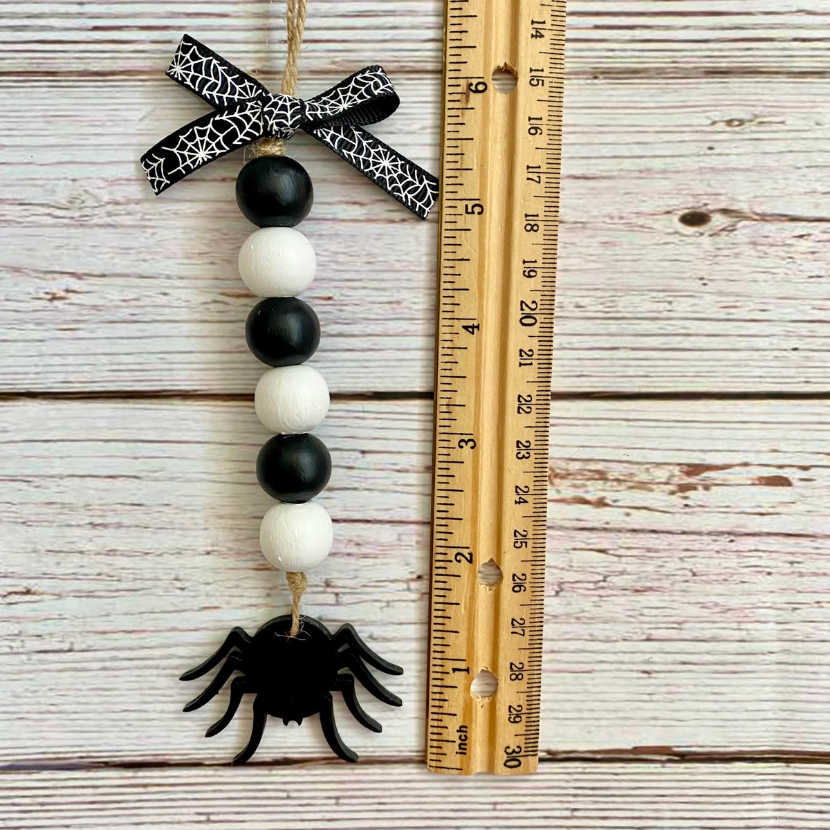 A Halloween bead garland ornament with a black spider, alternating black and white beads, and a black-and-white spiderweb ribbon bow, shown next to a wooden ruler on a whitewashed wood background.