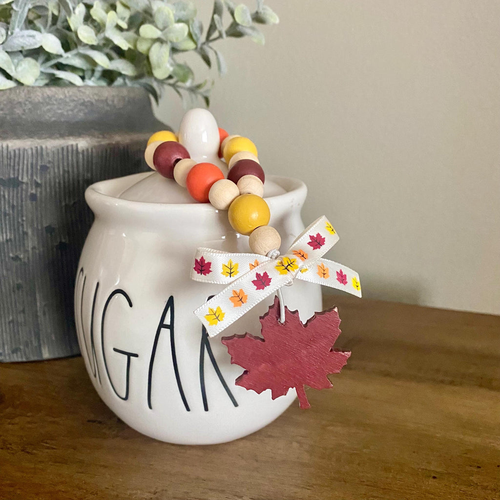 A white ceramic sugar bowl with 'SUGAR' printed in black on the front, styled on a wooden table with a beaded garland featuring wooden beads, a white ribbon with colorful maple leaves, and a red wooden maple leaf charm.