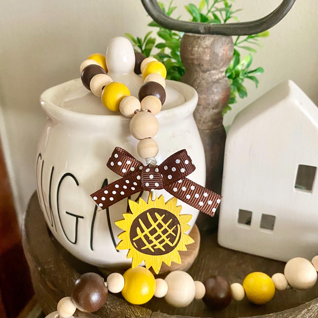 A beaded garland with natural wood, yellow, and dark brown beads, decorated with a brown polka-dot ribbon bow and a yellow sunflower charm, styled on a white ceramic canister.