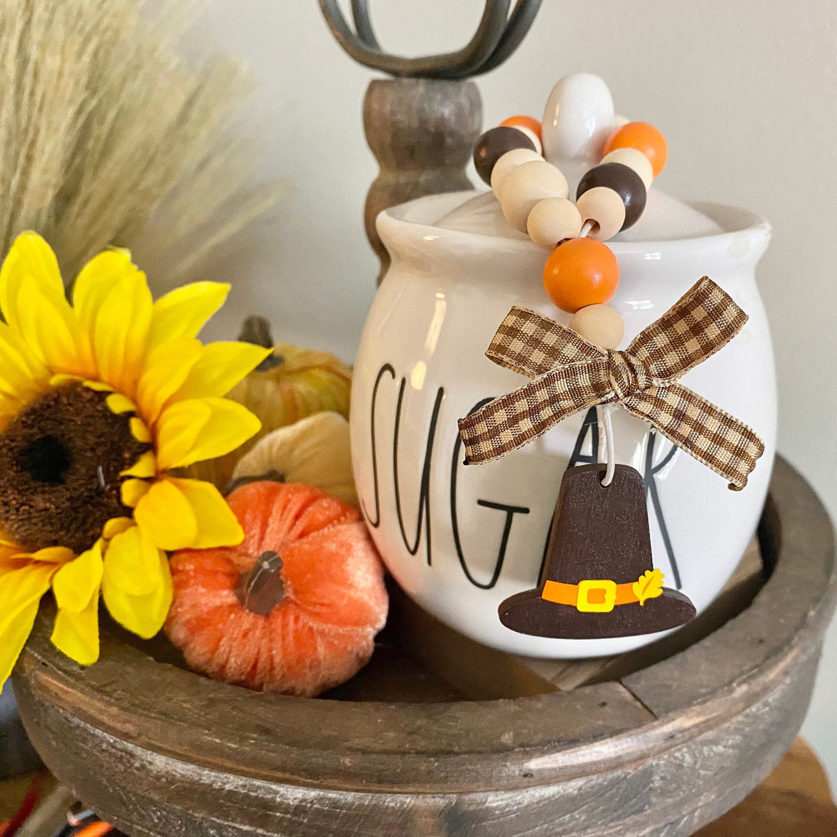A white ceramic canister with a lid, decorated with the word 'CUT' in black script, a brown plaid ribbon bow, a Pilgrim hat ornament, and a beaded garland, styled on a rustic wooden tray with a sunflower and small pumpkins.