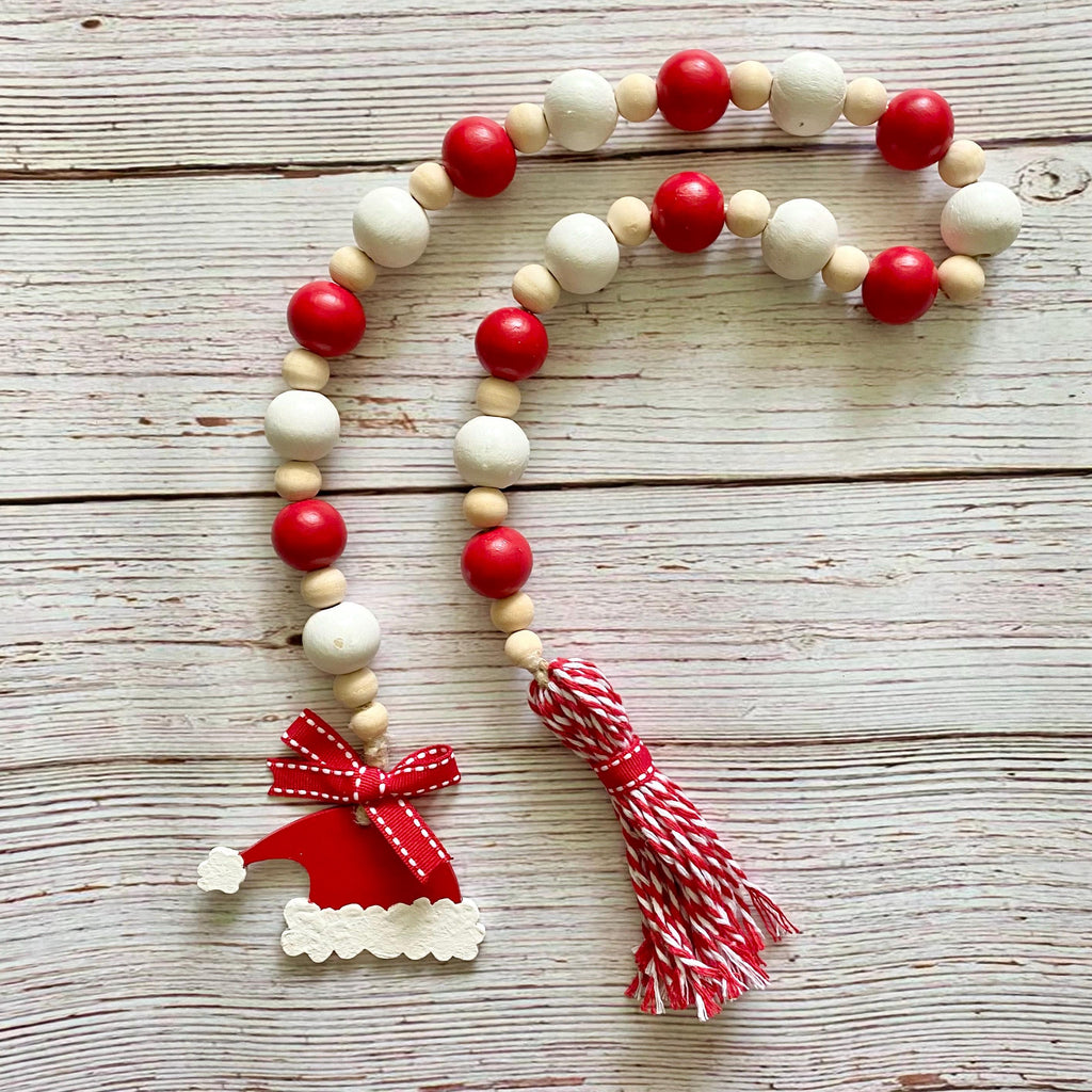 A Christmas-themed wooden bead garland with alternating red and natural white beads, a red Santa hat charm, and a red-and-white striped tassel, displayed on a rustic whitewashed wooden surface.