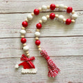 A Christmas-themed wooden bead garland with alternating red and natural white beads, a red Santa hat charm, and a red-and-white striped tassel, displayed on a rustic whitewashed wooden surface.