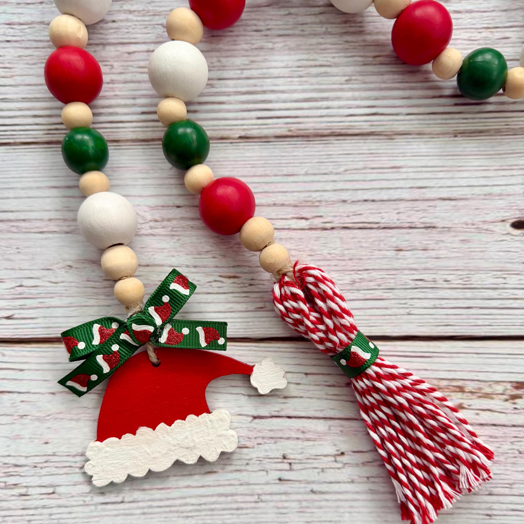 A Christmas-themed wooden bead garland necklace with red, green, and natural wood beads, a red and white patterned ribbon bow, a red Santa hat charm, and a red and white striped tassel, displayed on a whitewashed wooden background.