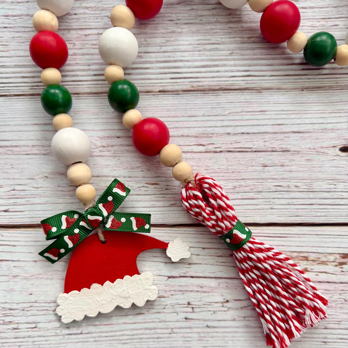 A Christmas-themed wooden bead garland necklace with red, green, and natural wood beads, a red and white patterned ribbon bow, a red Santa hat charm, and a red and white striped tassel, displayed on a whitewashed wooden background.