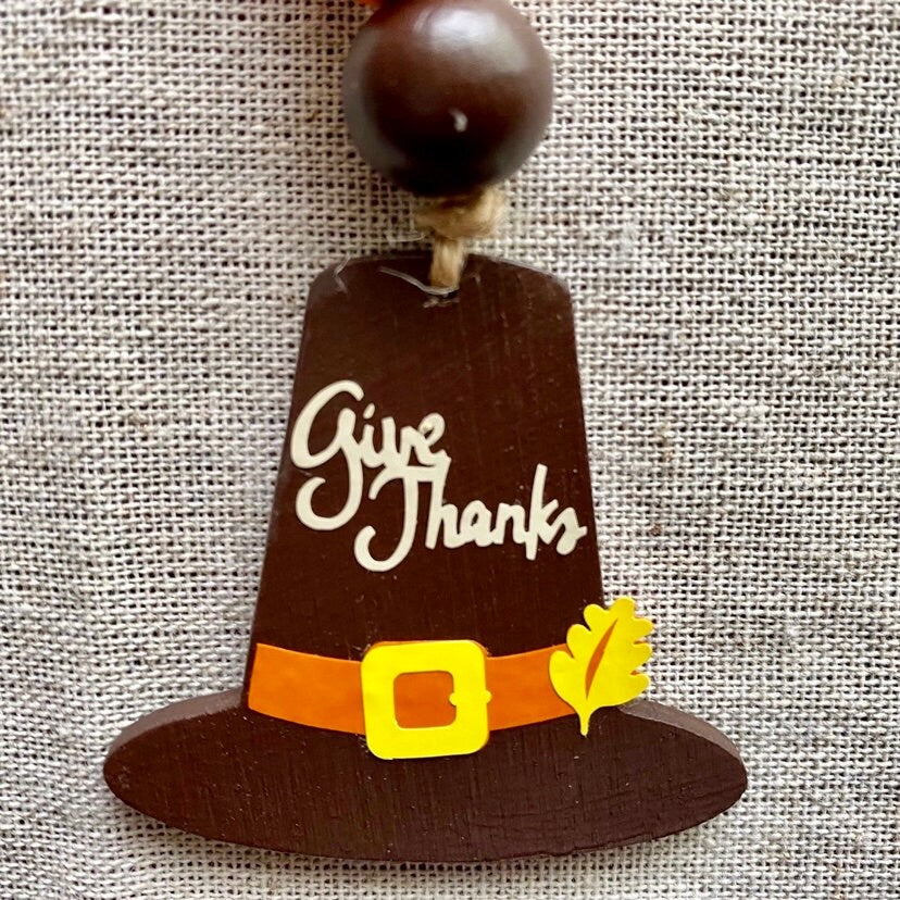 A wooden Thanksgiving ornament shaped like a pilgrim hat, with 'Give Thanks' written in white script, an orange band, a yellow buckle, and a small yellow leaf, hanging against a beige burlap background.