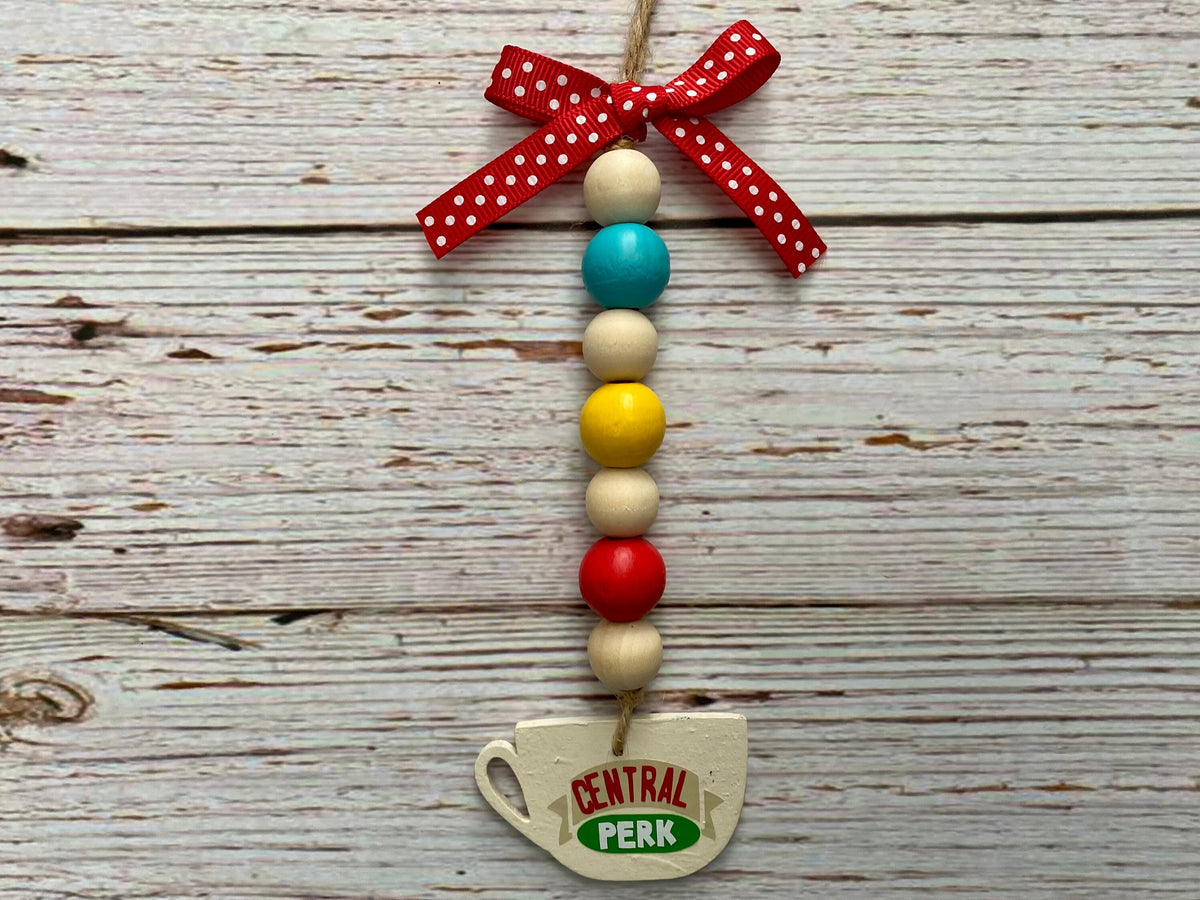 A decorative hanging ornament shaped like a coffee cup with the words 'CENTRAL Perk' on it, suspended from a string with colorful wooden beads and a red polka-dot ribbon bow, set against a rustic whitewashed wood background.