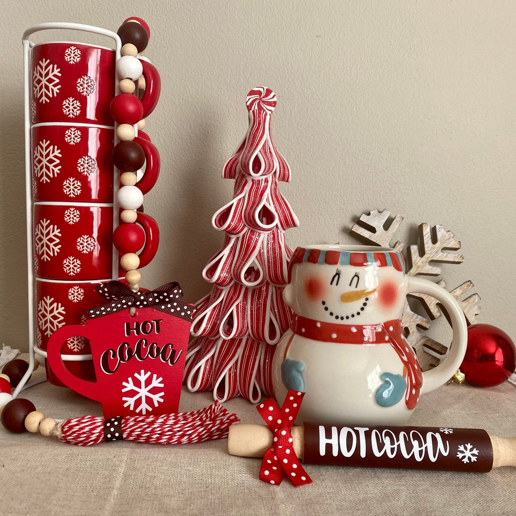 A holiday-themed hot cocoa gift set arranged on a tabletop against a plain beige wall. The set includes a tall stack of red cups with white snowflakes, a red and white striped decorative tree, a snowman mug, a red heart-shaped tag reading 'HOT COCOA', a red-and-white striped candy cane, a red-and-white striped rolling pin, and a red ornament.