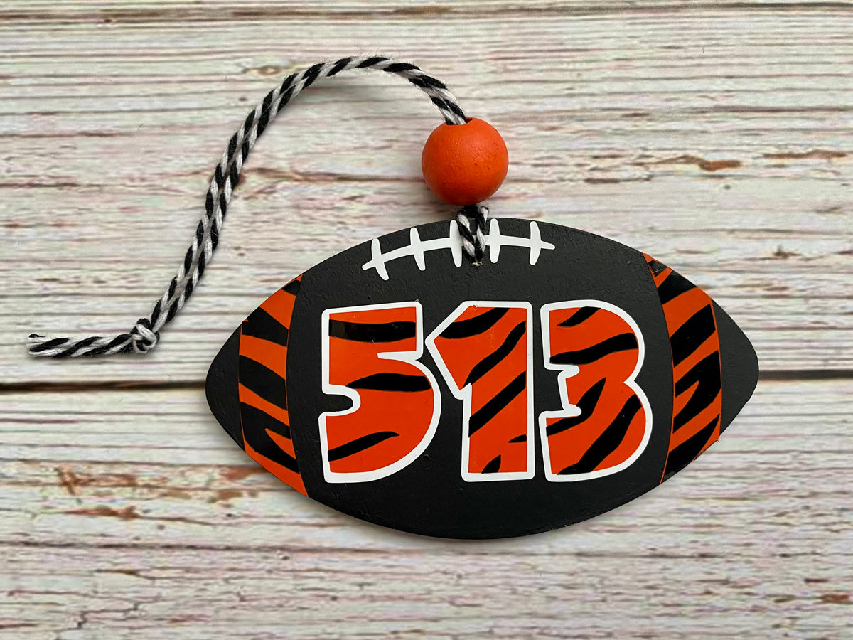 A football-shaped wooden ornament with orange and black tiger-stripe patterns, white laces, and the number 513 in orange with white outlines. It hangs from a black-and-white string with an orange bead, set against a rustic light wood background.