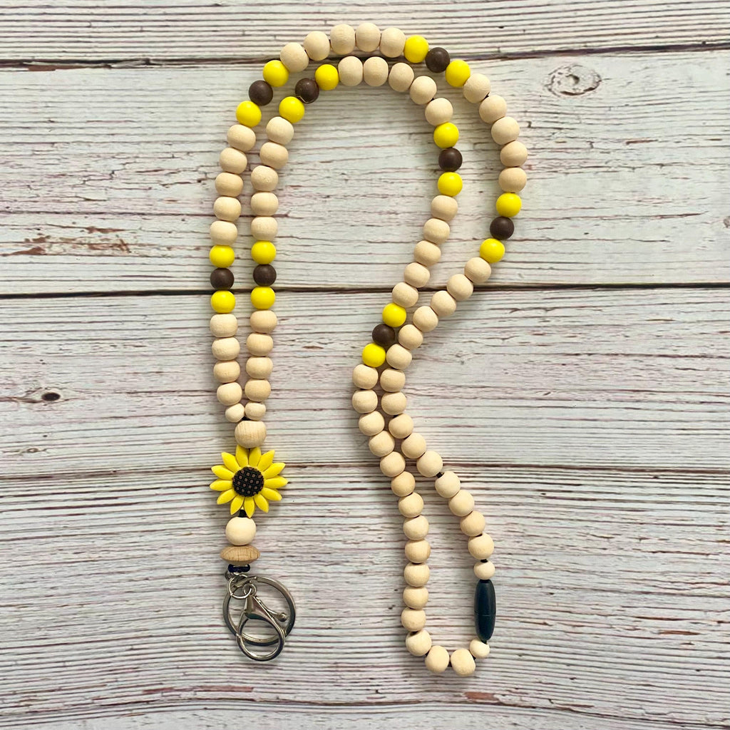 A wooden bead lanyard or keychain necklace with natural beige beads, bright yellow beads, dark brown beads, and a yellow sunflower charm, laid flat on a whitewashed wooden surface.