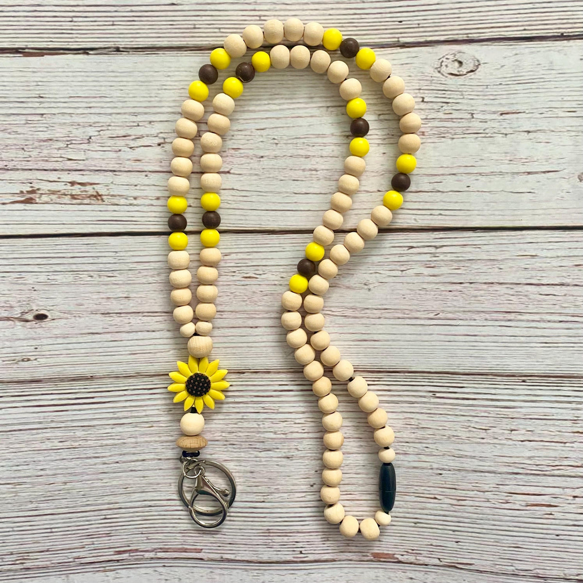 A wooden bead lanyard or keychain necklace with natural beige beads, bright yellow beads, dark brown beads, and a yellow sunflower charm, laid flat on a whitewashed wooden surface.