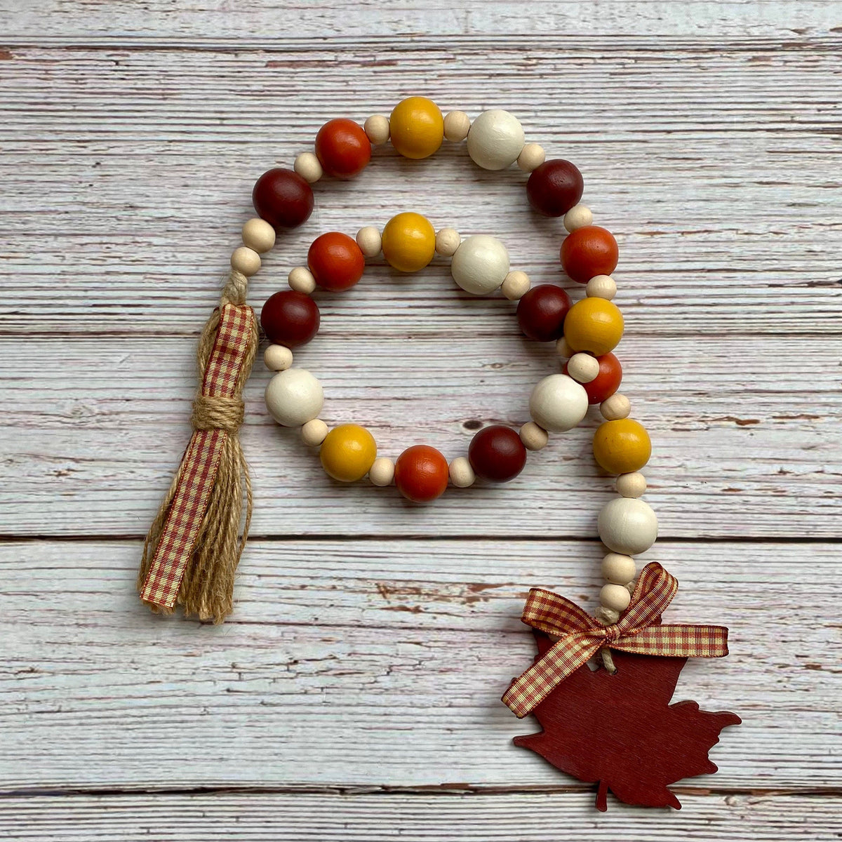 A wooden bead garland with autumn colors, featuring red, orange, yellow, and cream beads, a brown gingham tassel, and a red maple leaf charm tied with a matching ribbon, displayed on a rustic white wood background.