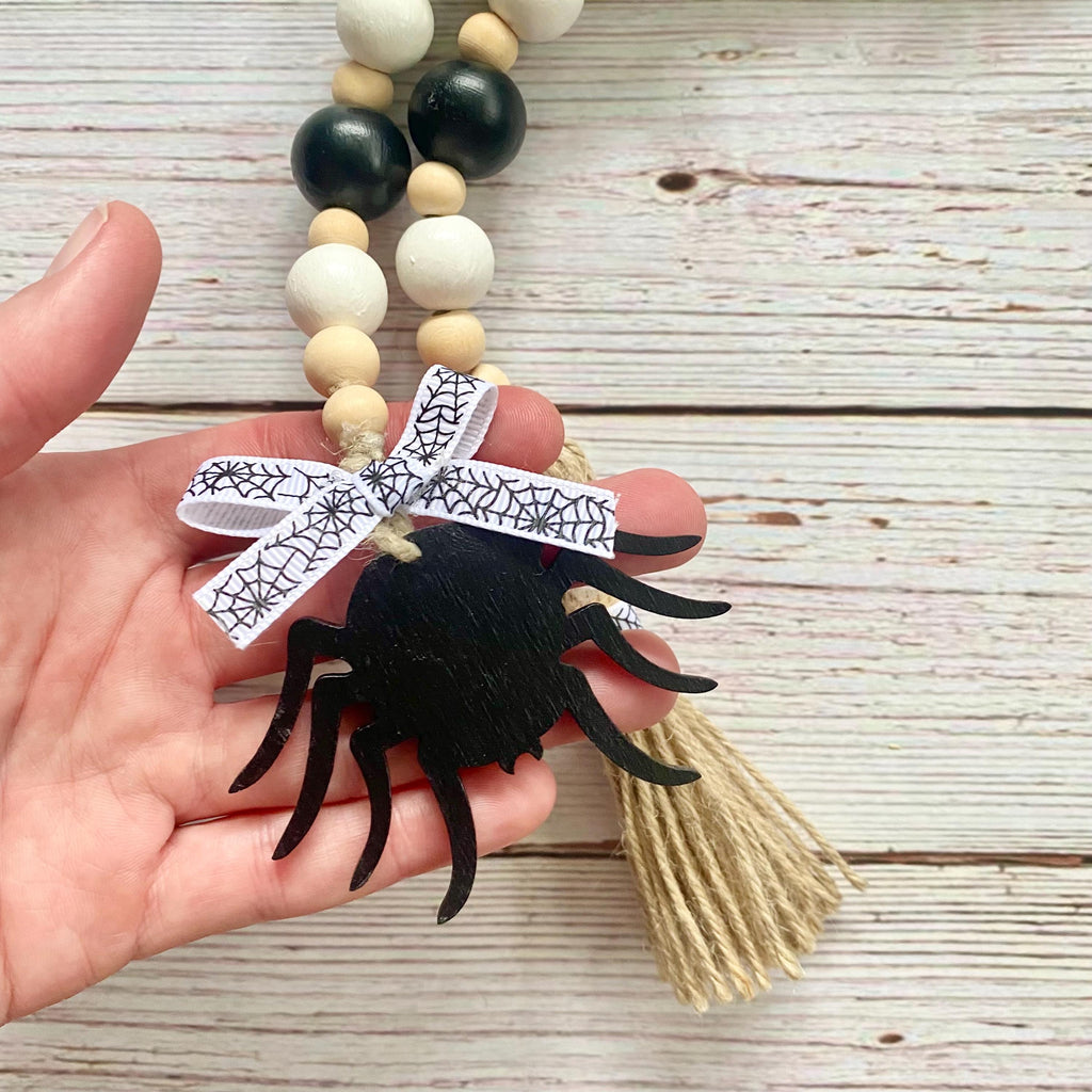 A hand holding a wooden bead garland with a black spider ornament, a white spiderweb ribbon bow, and a beige tassel, set against a rustic white-washed wood background.