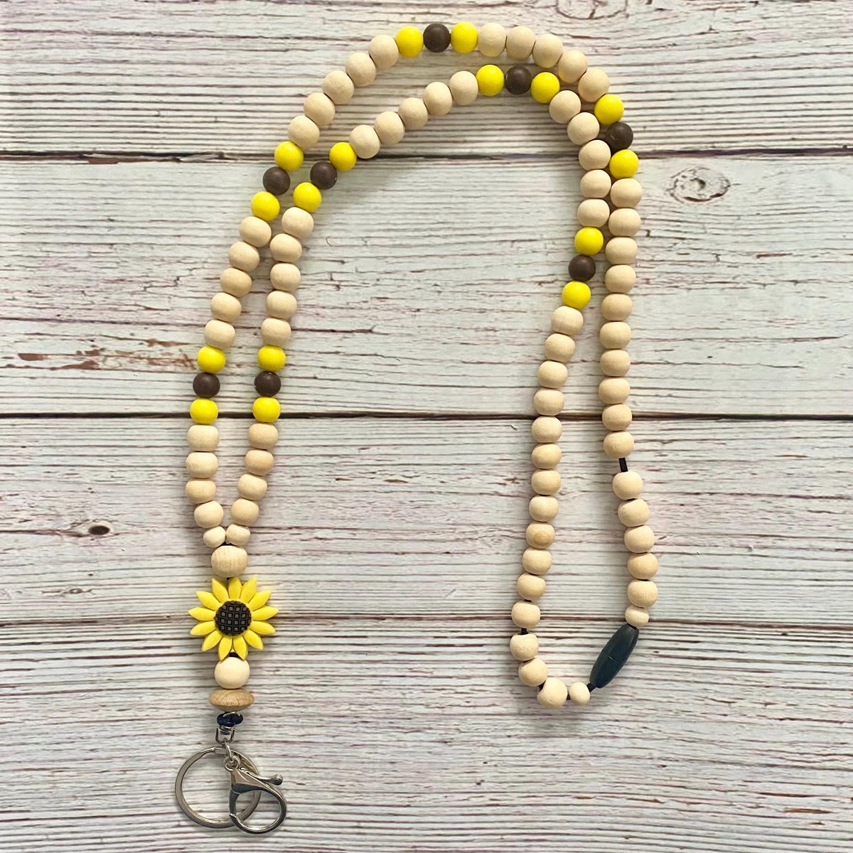 A beaded necklace or car keychain strap made of natural wood beads with bright yellow and dark brown accent beads, a yellow sunflower charm, and a metal key ring clasp, laid flat on a whitewashed wooden surface.